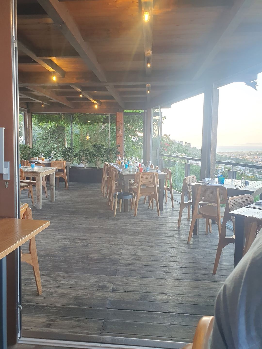An elevated dining terrace with wooden tables and chairs overlooking a coastal landscape, featuring floor-to-ceiling windows and warm ambient lighting.