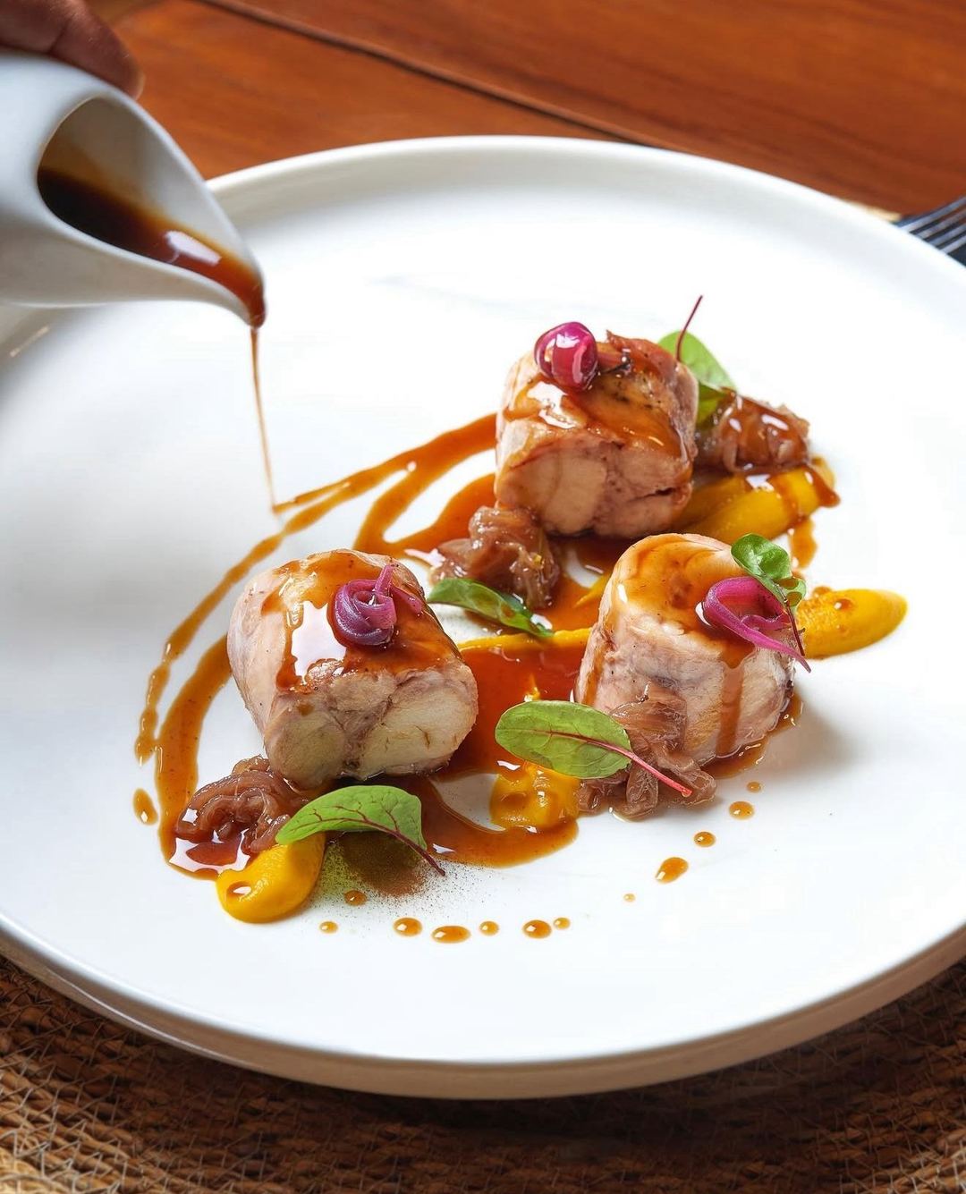 A plated dish of seared scallops with caramelized sauce, yellow vegetables, and microgreens garnish being finished with a brown reduction sauce.