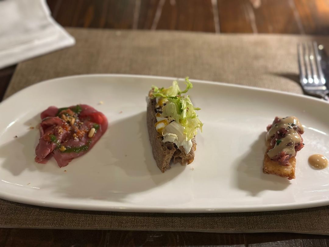 An elegantly plated charcuterie and appetizer course featuring three refined bites with cured meat, topped vegetables, and garnishes on a white plate.