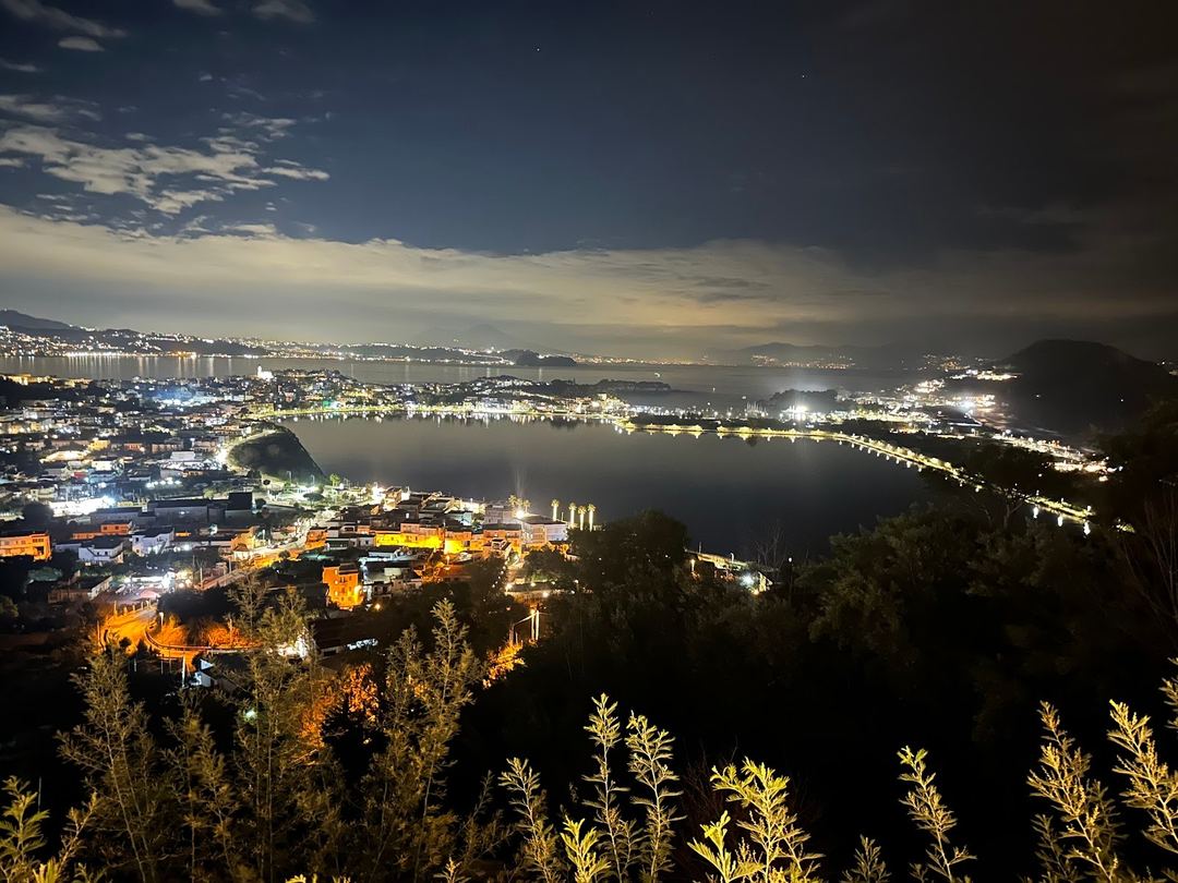 Nighttime panoramic view of a coastal bay with illuminated waterfront buildings and surrounding hills.