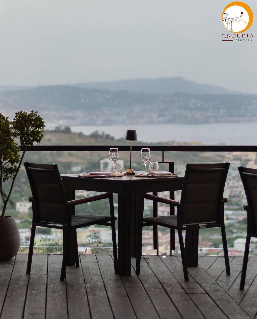 An elegantly set dining table on a terrace overlooks a scenic lake landscape with distant mountains and coastal settlements.
