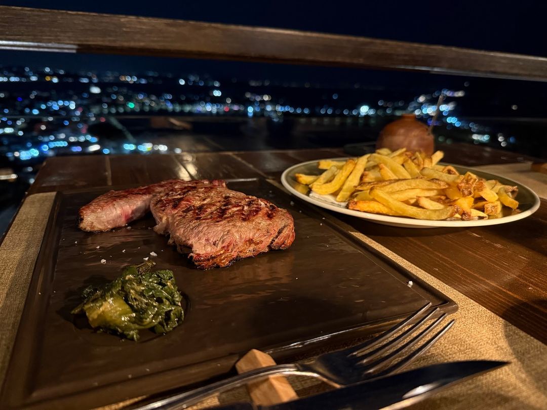 Grilled steaks with chimichurri sauce and golden french fries served on a wooden board overlooking a night cityscape.