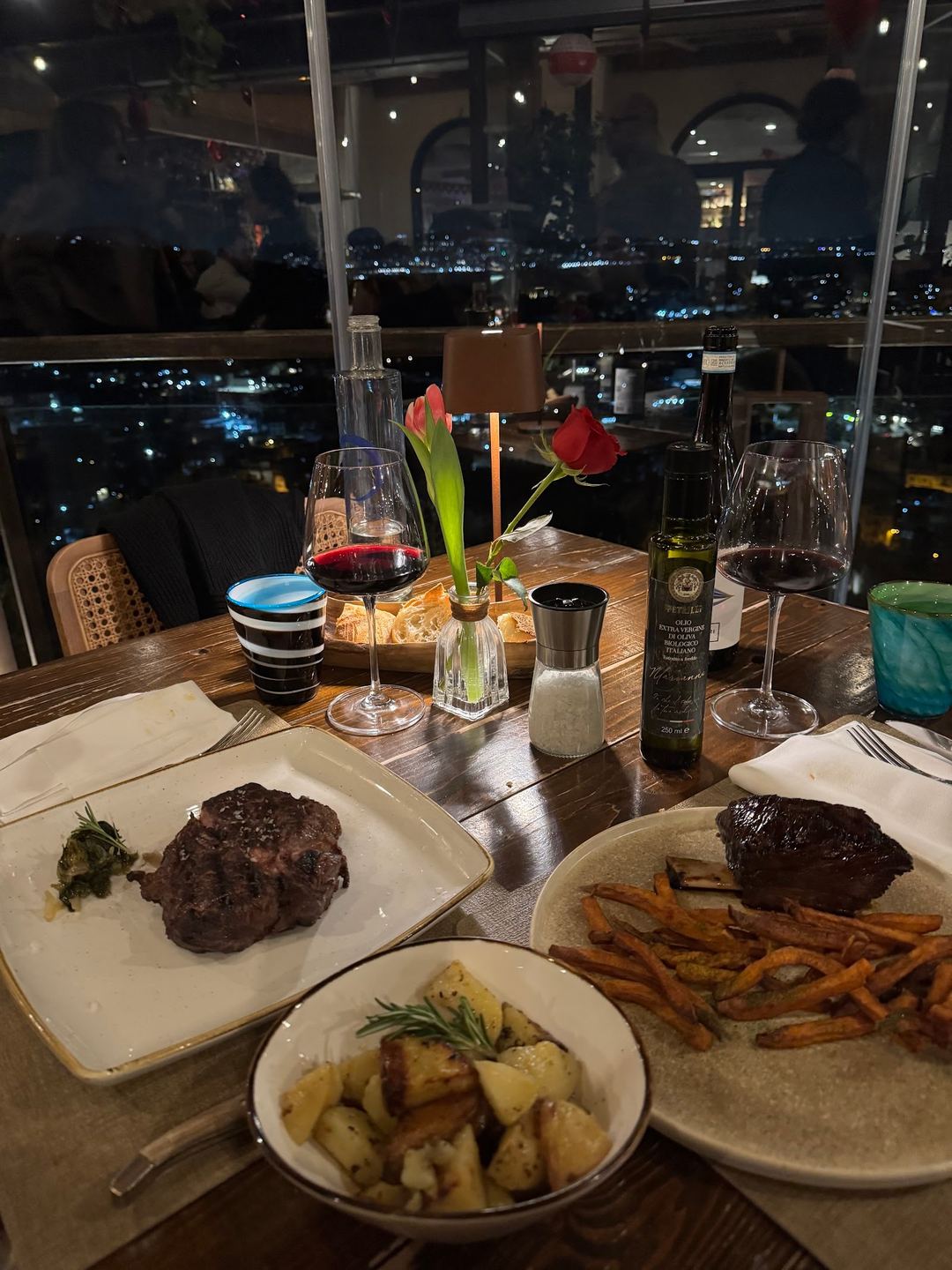 An elegant dinner table with grilled steaks, fries, roasted potatoes, and red wine overlooking a nighttime city skyline.