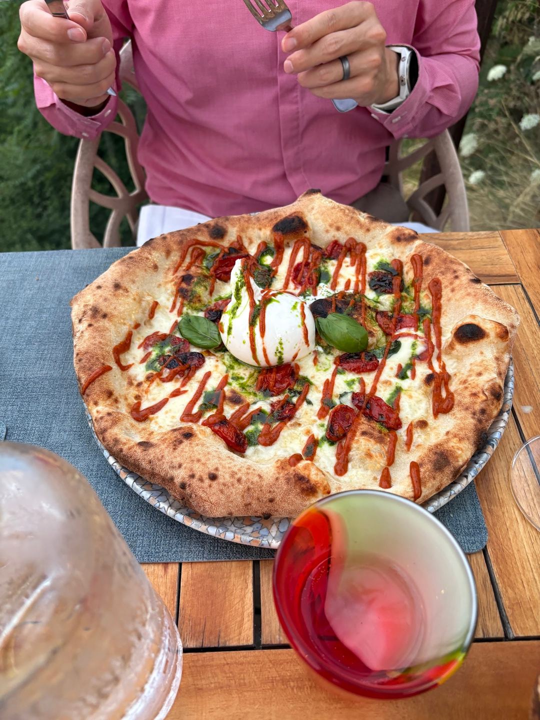 A wood-fired pizza topped with burrata cheese, tomato sauce, fresh basil, and crispy pancetta, served outdoors at a restaurant table.